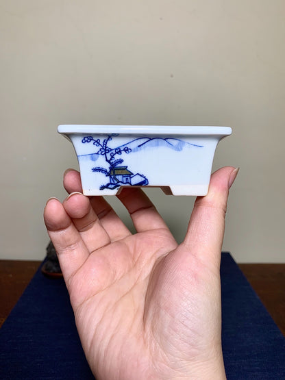 A hand holds a small white ceramic pot with blue painted landscape and tree design.