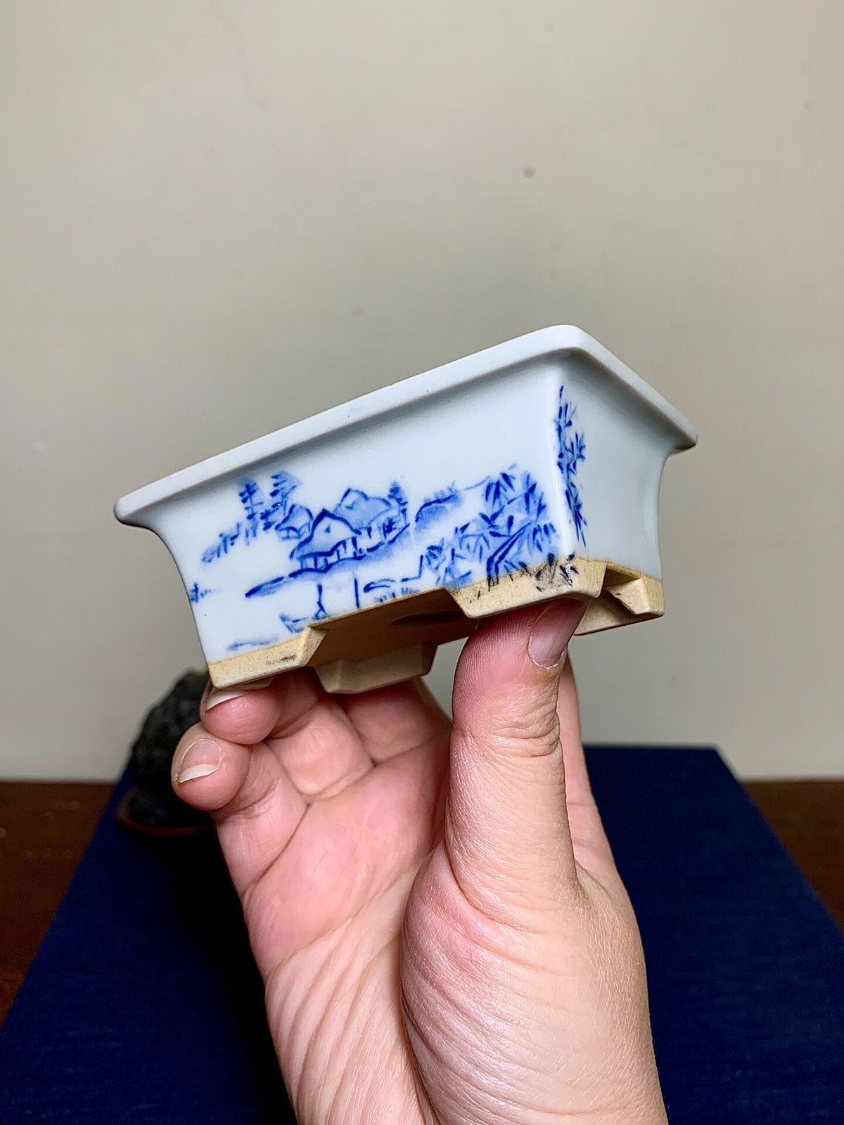 A hand holds a white rectangular pot with blue landscape painting and a glazed finish.