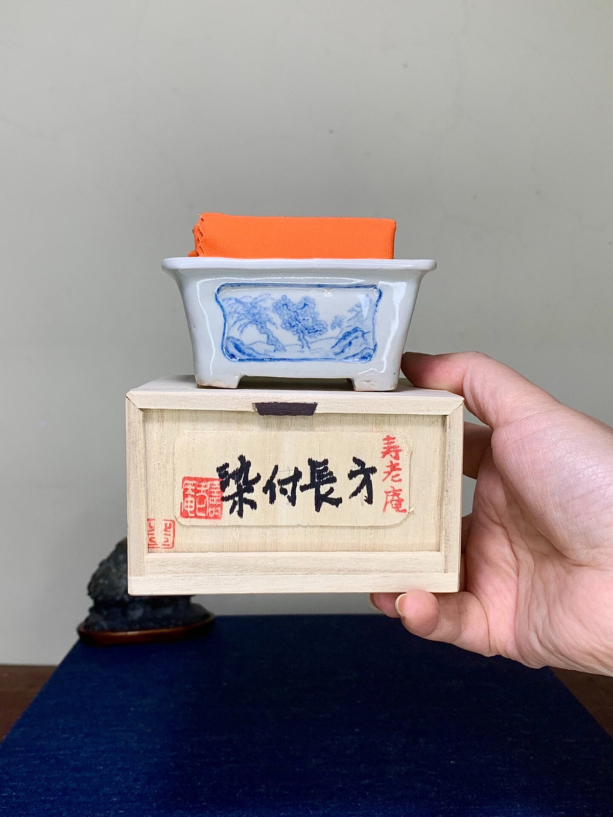Hand holds white glazed pot with blue landscape, atop wooden box with black and red characters.