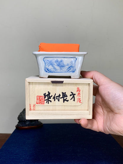 Hand holds white glazed pot with blue landscape, atop wooden box with black and red characters.