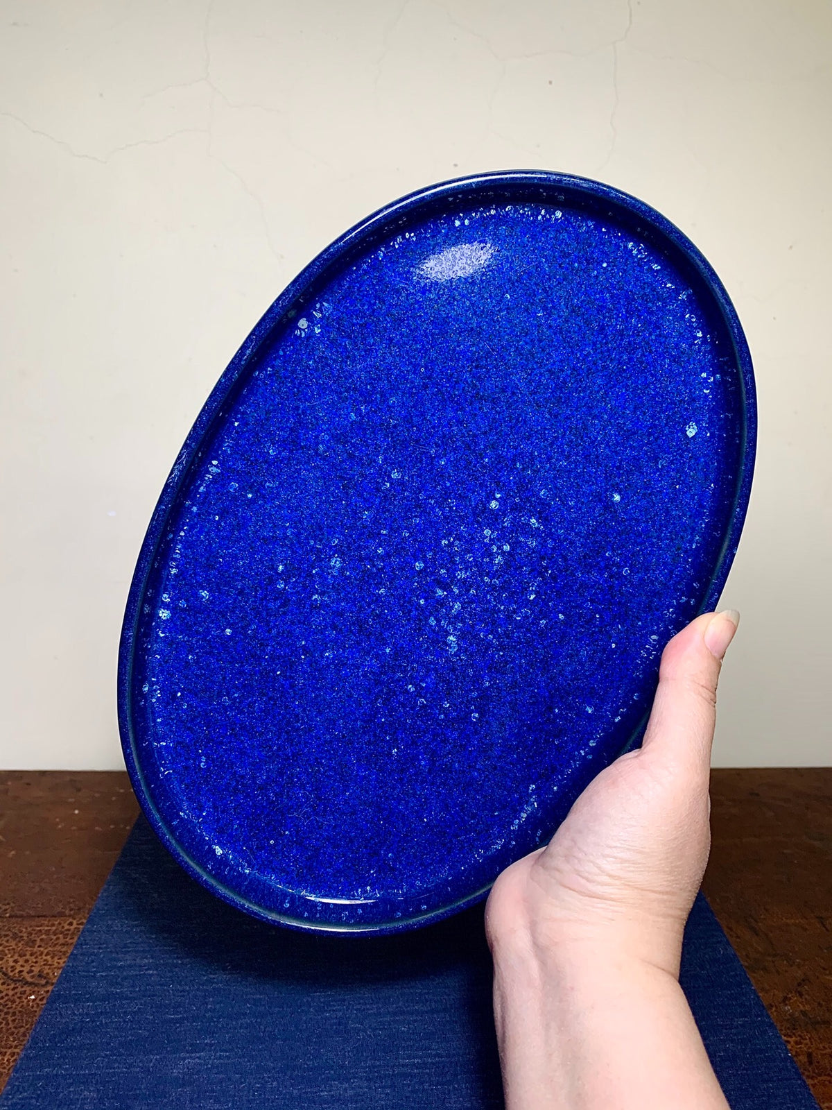 A hand holds an oval, glossy cobalt blue ceramic tray with white speckles.