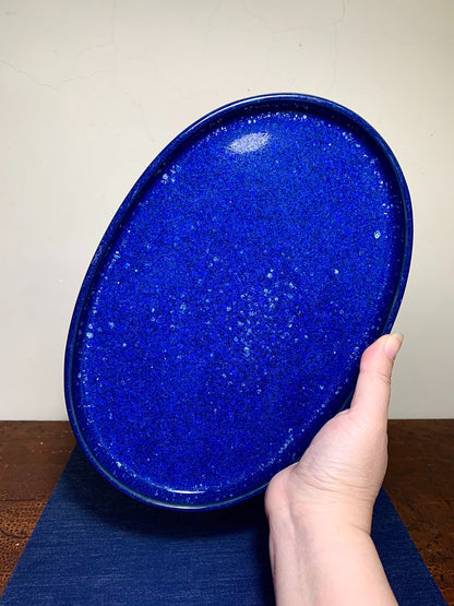A hand holds an oval, glossy cobalt blue ceramic tray with white speckles.