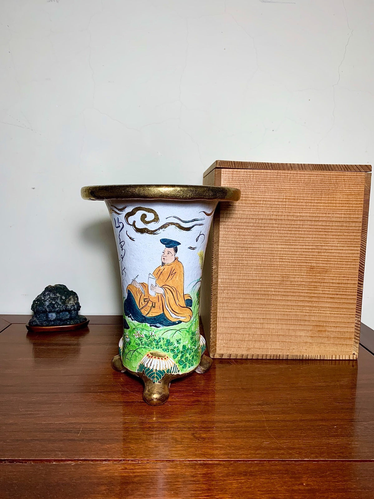 White glazed pot with painted figure, gold rim and feet, beside wooden box.