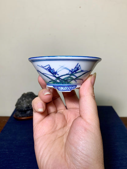 A hand holds a small white bowl with blue and green painted grass and insect motifs.