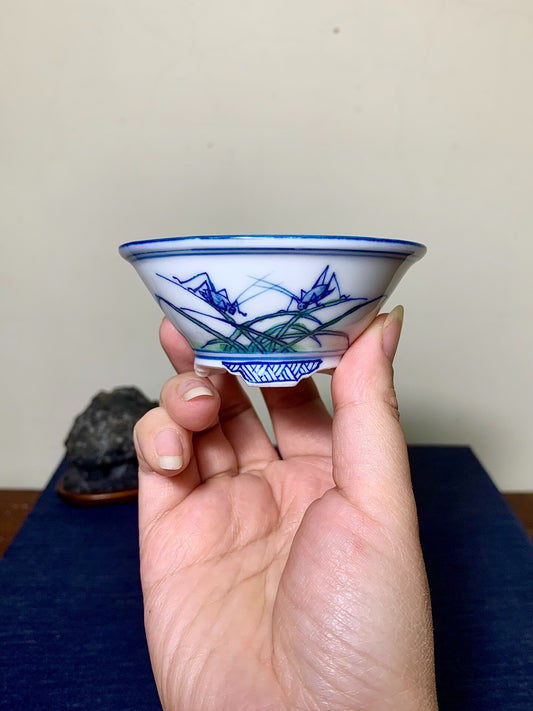 A hand holds a small white bowl with blue and green painted grass and insect motifs.