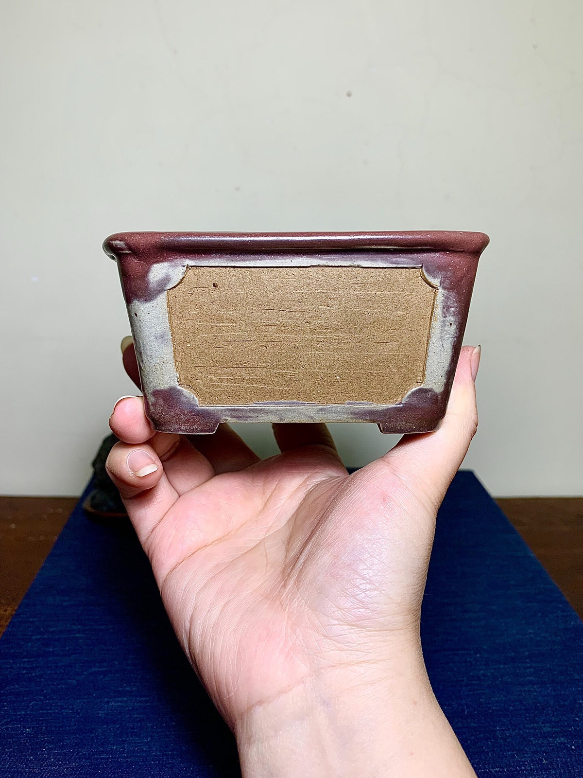 A hand holds a small, rectangular, reddish-brown glazed pot with a beige unglazed interior.