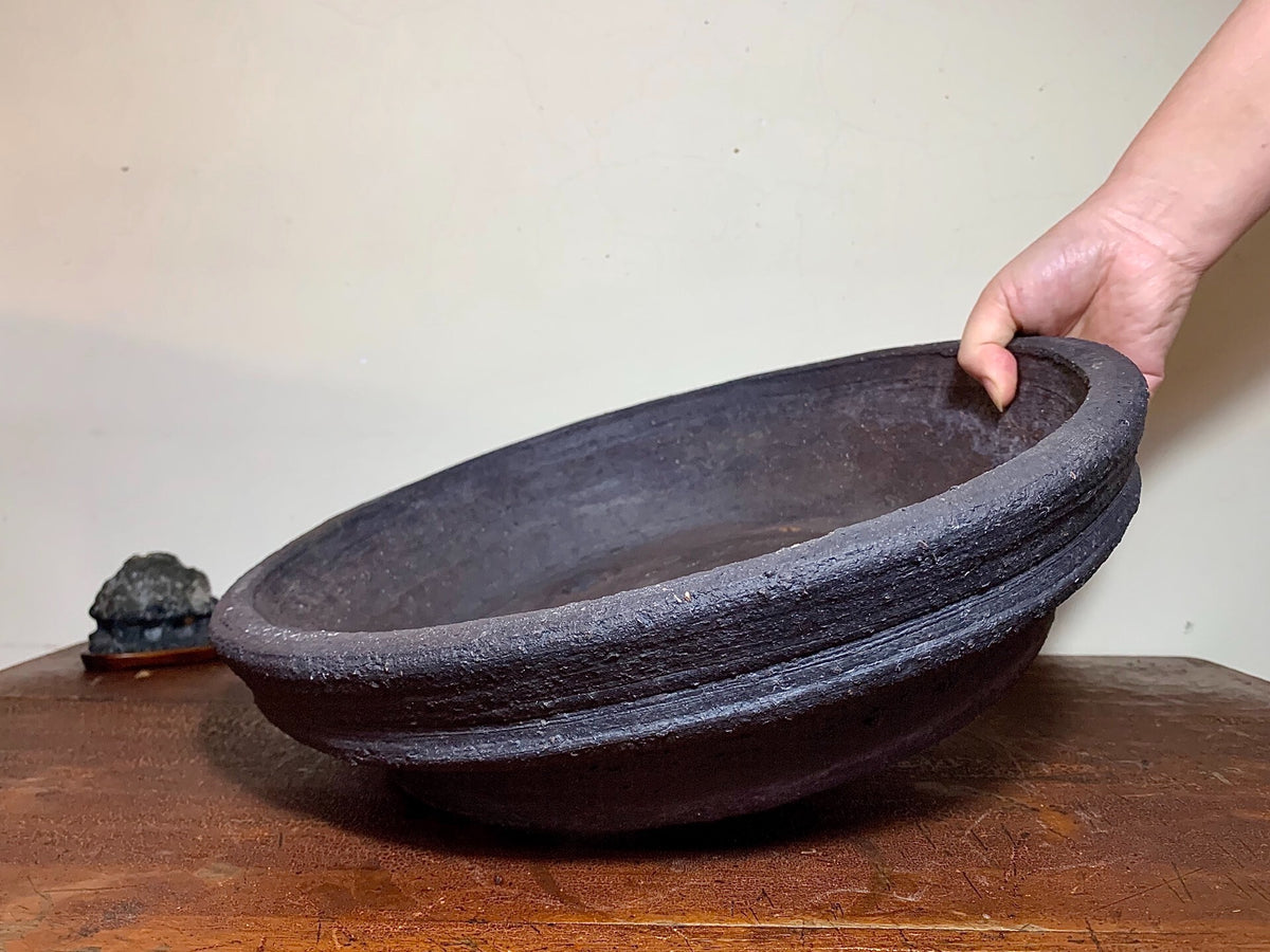 A hand holds a large, dark, unglazed ceramic pot with a thick, textured rim.