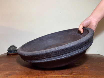 A hand holds a large, dark, unglazed ceramic pot with a thick, textured rim.