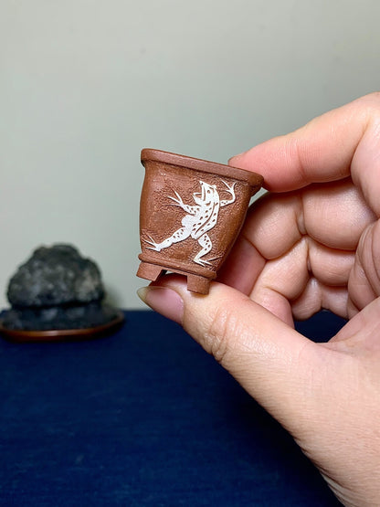 A hand holds a small, brown, unglazed ceramic pot with a white frog design.