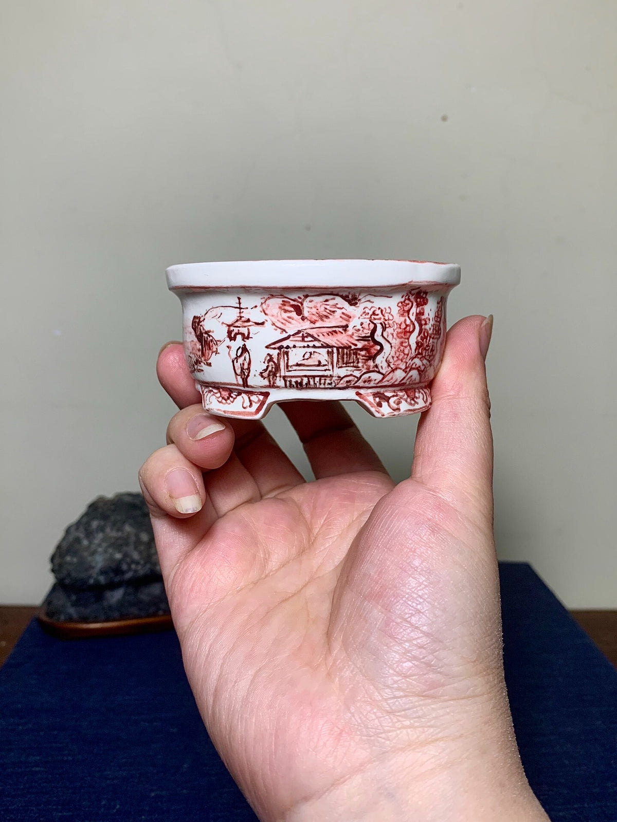 A hand holds a white ceramic pot with red painted landscape scenes and a slightly chipped rim.