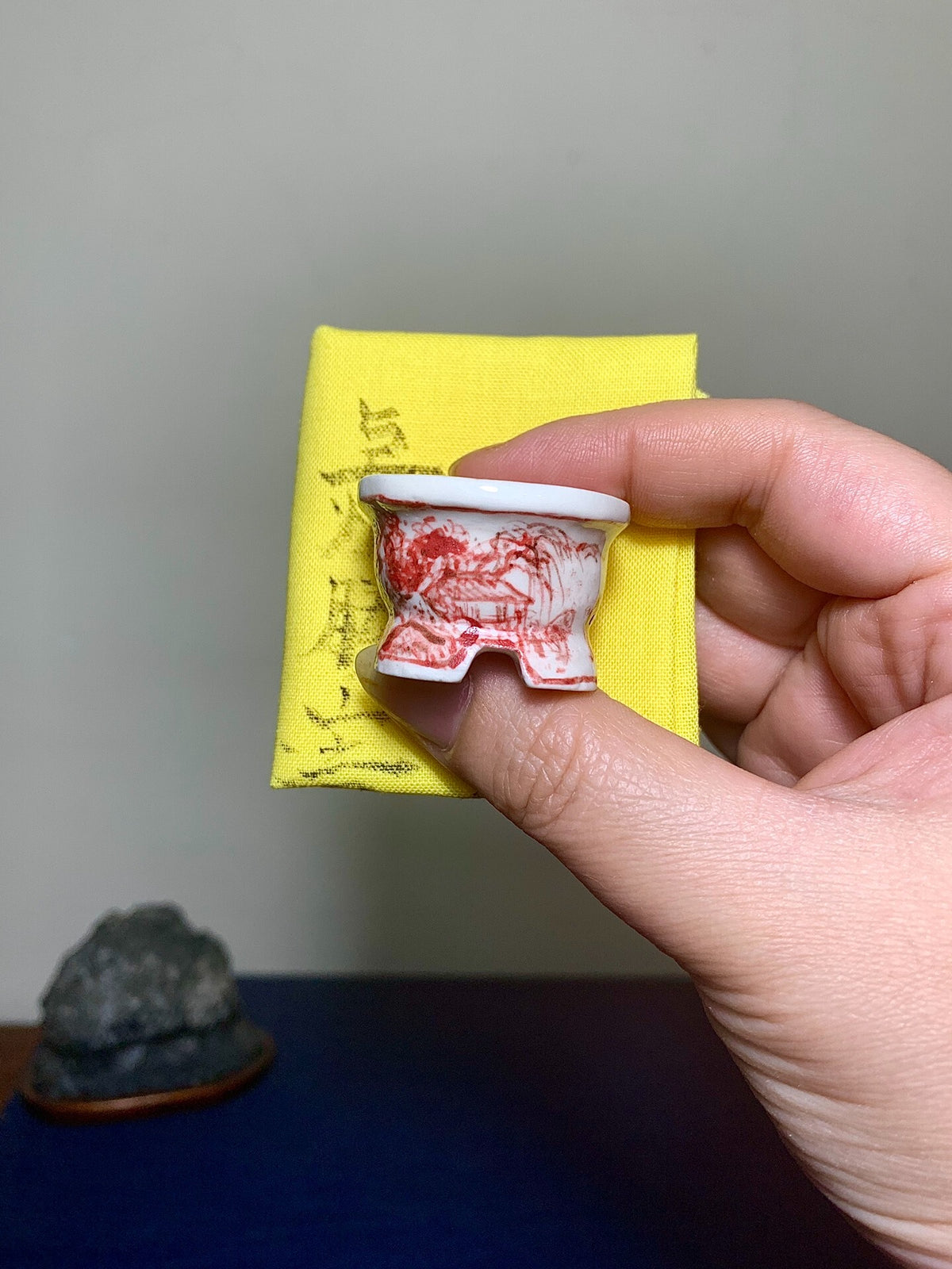 A hand holds a small white ceramic pot with red painted landscape decoration.