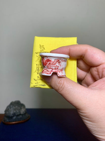 A hand holds a small white ceramic pot with red painted landscape decoration.