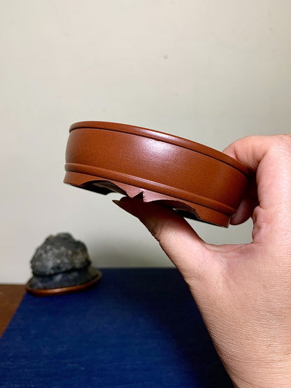 A hand holds a glossy, reddish-brown ceramic pot base with a scalloped rim and three feet.