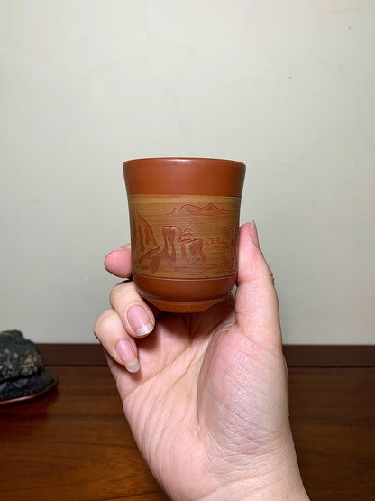 A hand holds a small, brown ceramic cup with a glazed, landscape scene etched around its middle.
