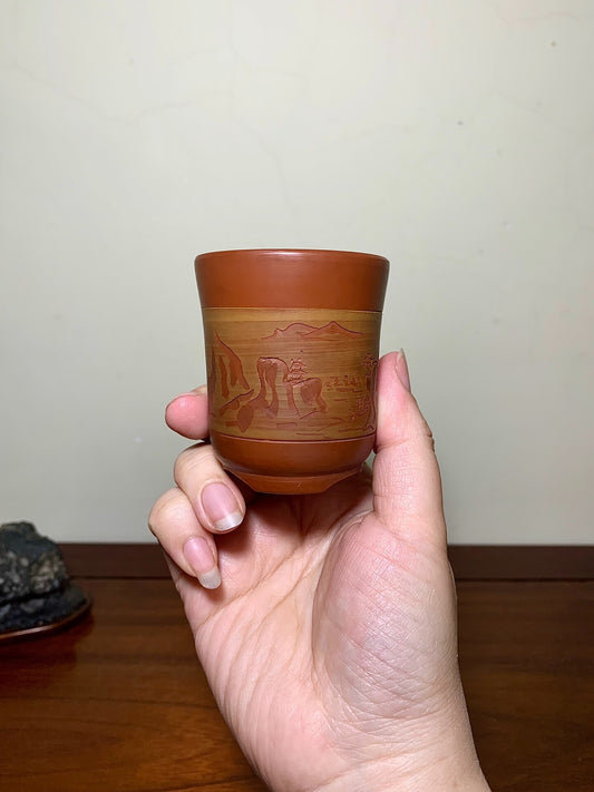 A hand holds a small, brown ceramic cup with a glazed, landscape scene etched around its middle.