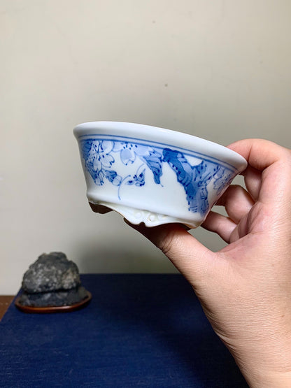 A hand holds a white glazed ceramic pot with blue floral and leaf patterns.