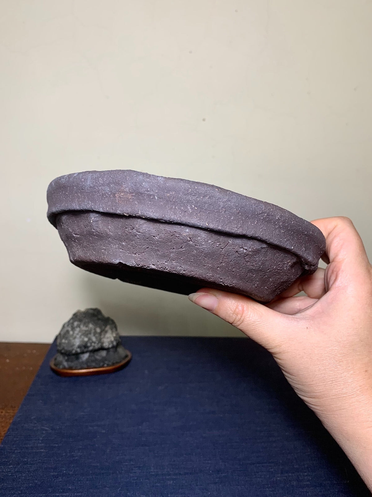 A hand holds a dark, unglazed, roughly textured round pot with a thick rim.