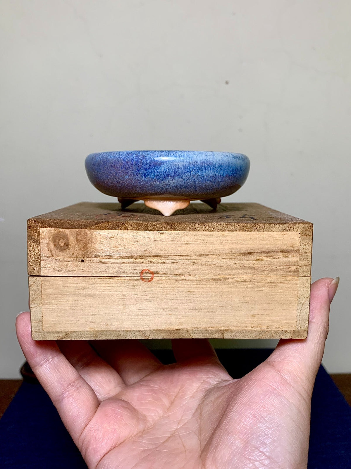 A hand holds a wooden box with a small, blue-glazed ceramic pot on top.