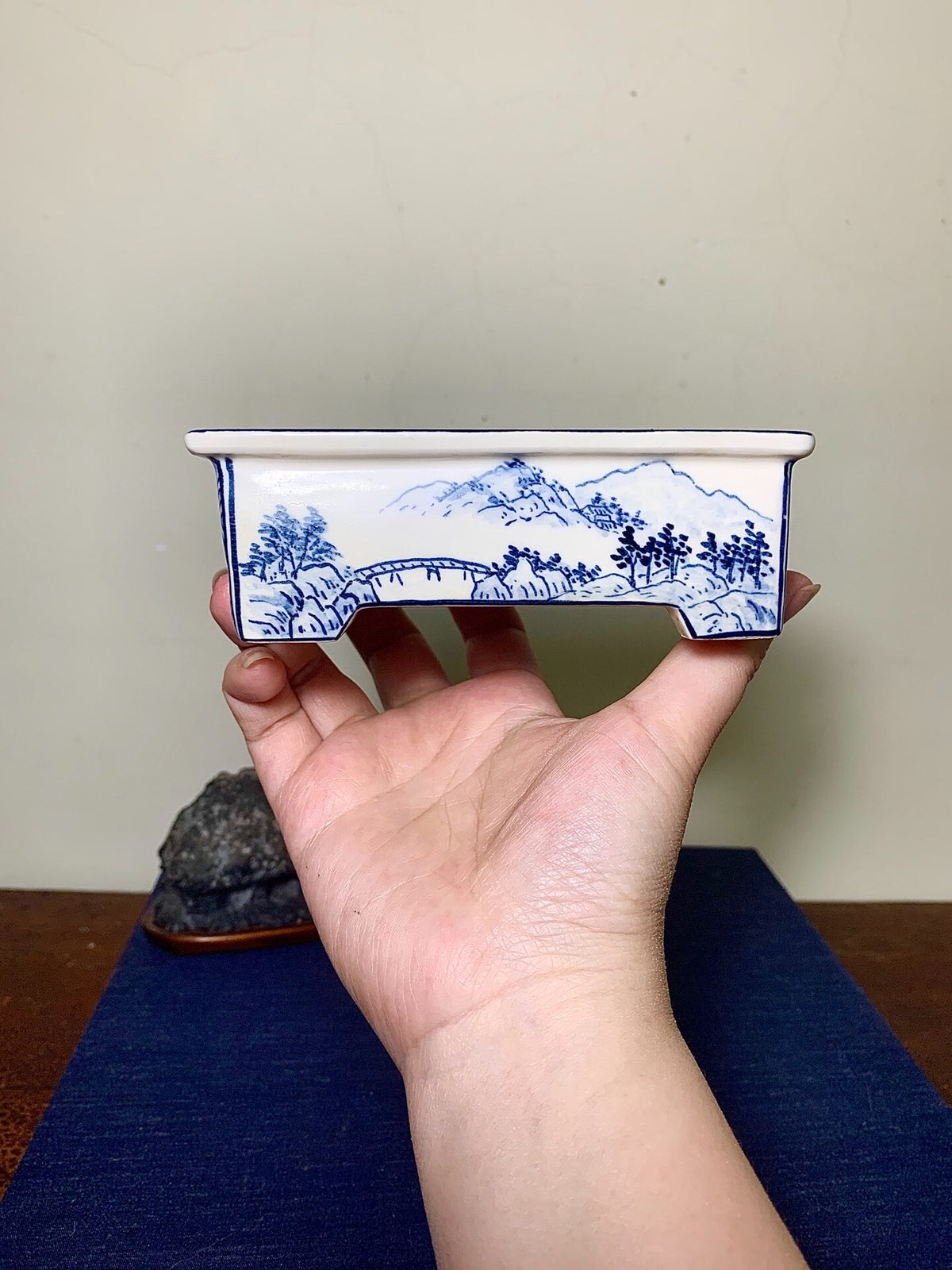 A hand holds a small, white, rectangular ceramic pot with blue landscape painting.