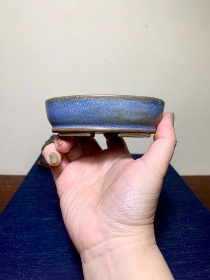 A hand holds a small, shallow, blue-glazed ceramic pot with a speckled finish and unglazed rim.