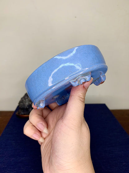 A hand holds a small, light blue glazed pot with white crackle patterns and four footed supports.