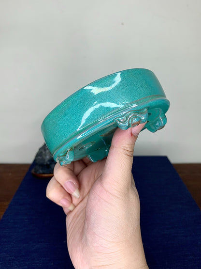 A hand holds a glossy teal glazed pot with white crackle patterns and scalloped foot.