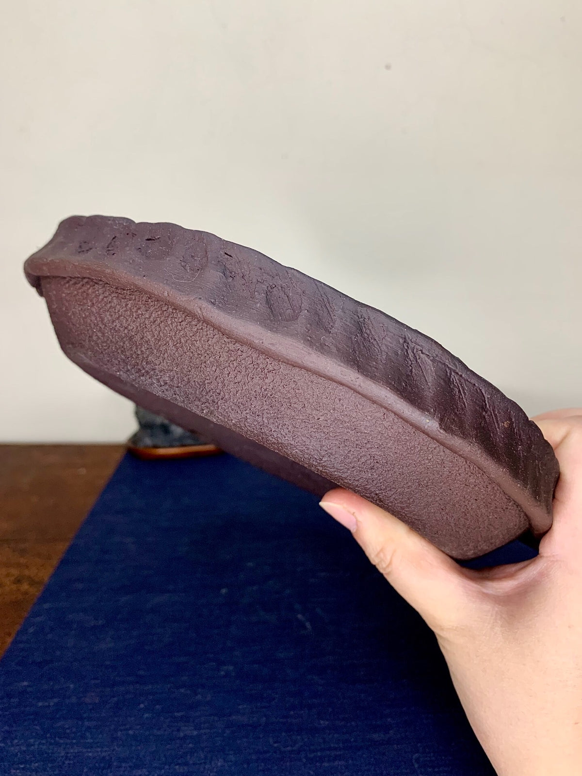 A hand holds a dark brown, unglazed ceramic pot with a textured, slightly uneven rim.