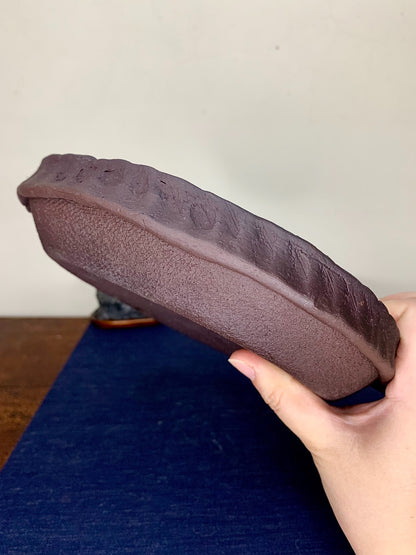 A hand holds a dark brown, unglazed ceramic pot with a textured, slightly uneven rim.