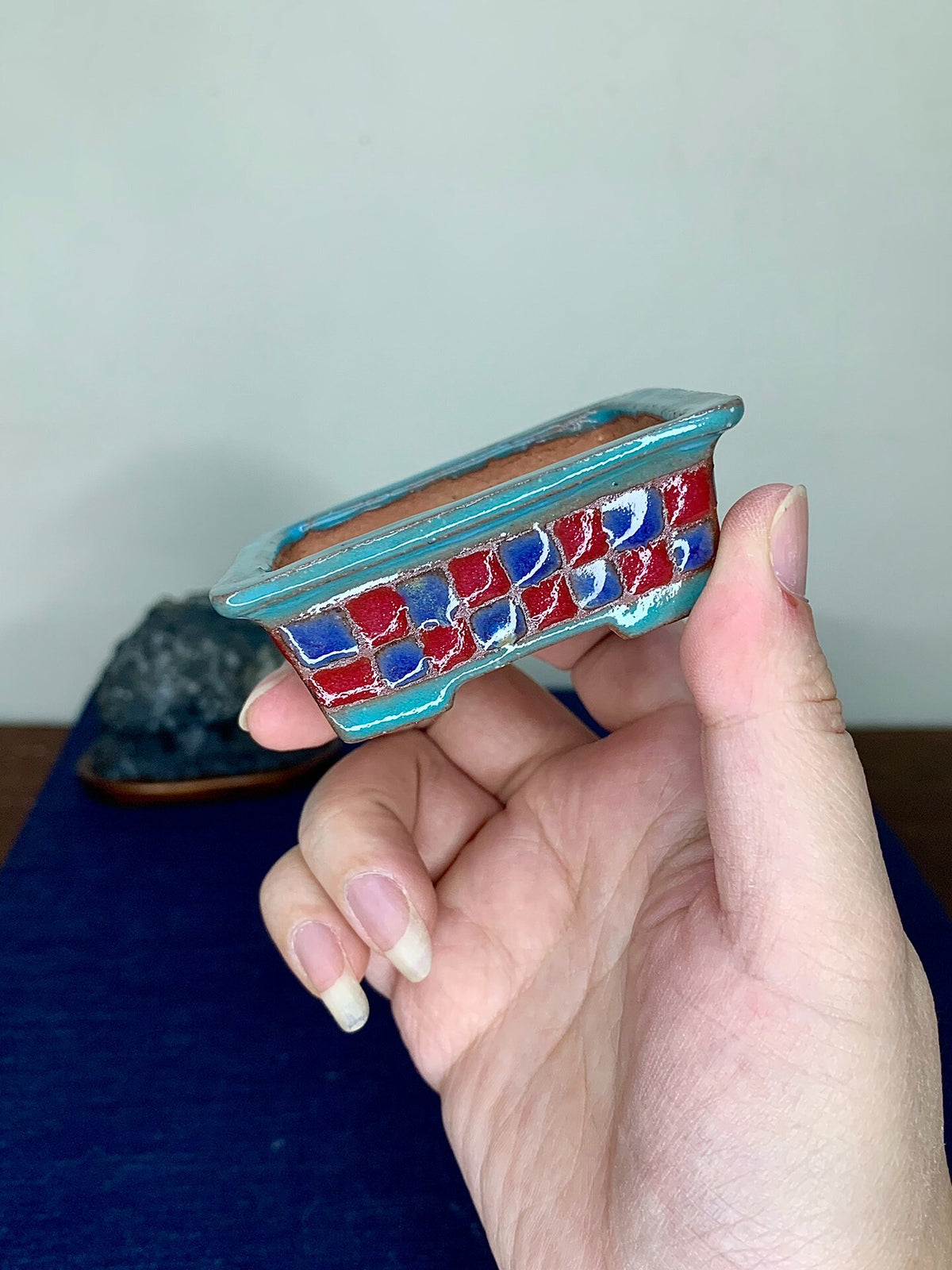 A hand holds a small rectangular pot with a turquoise glazed rim and red-blue mosaic patterned sides.