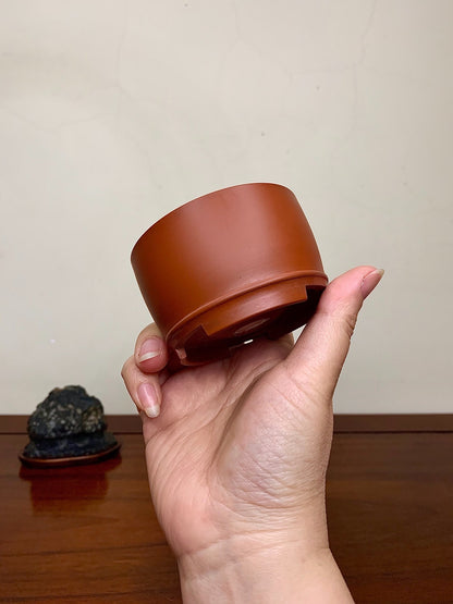 A hand holds a matte, terracotta-colored, cylindrical pot with a flared rim and unglazed base.