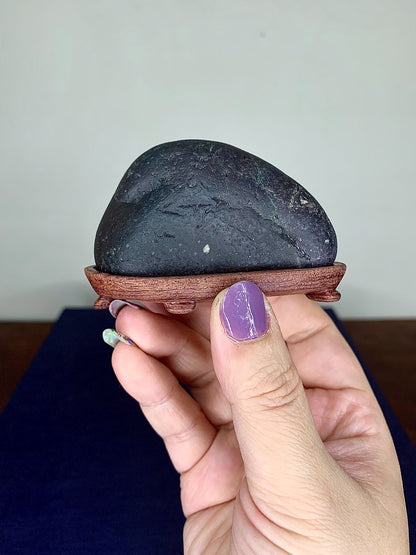 A hand holds a smooth, dark grey, unglazed stone on a small wooden stand.