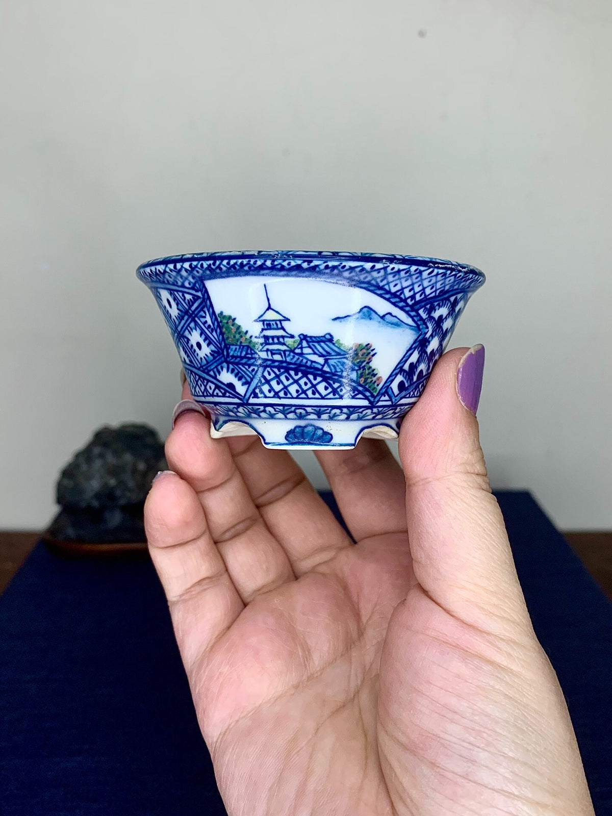 A hand holds a small blue and white ceramic pot with a painted landscape scene.