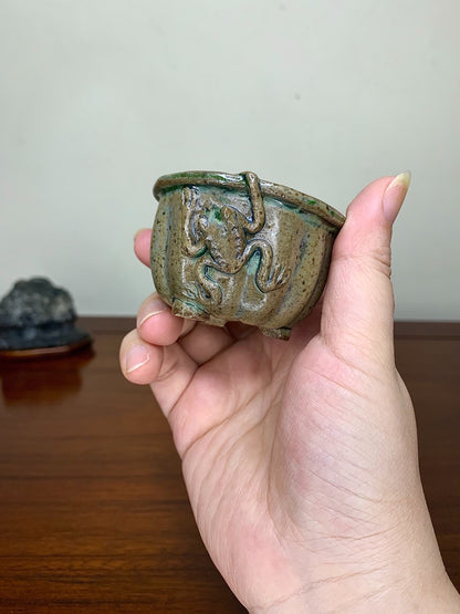 A hand holds a small, glazed ceramic pot with a dragon relief and mossy green glaze.