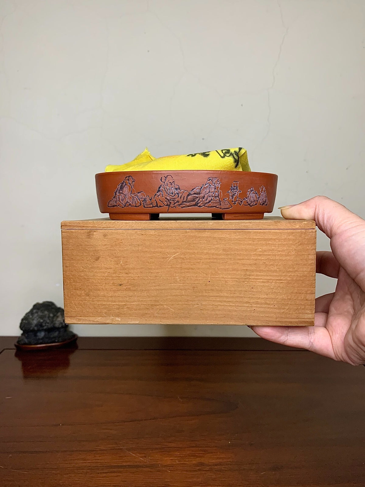 A hand holds a small, reddish-brown glazed pot with carved mountain scenes, resting on a wooden block.