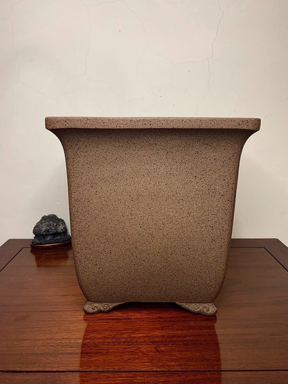 A brown speckled unglazed rectangular pot with four carved feet sits on a wooden table.