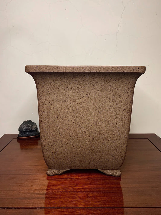 A brown speckled unglazed rectangular pot with four carved feet sits on a wooden table.