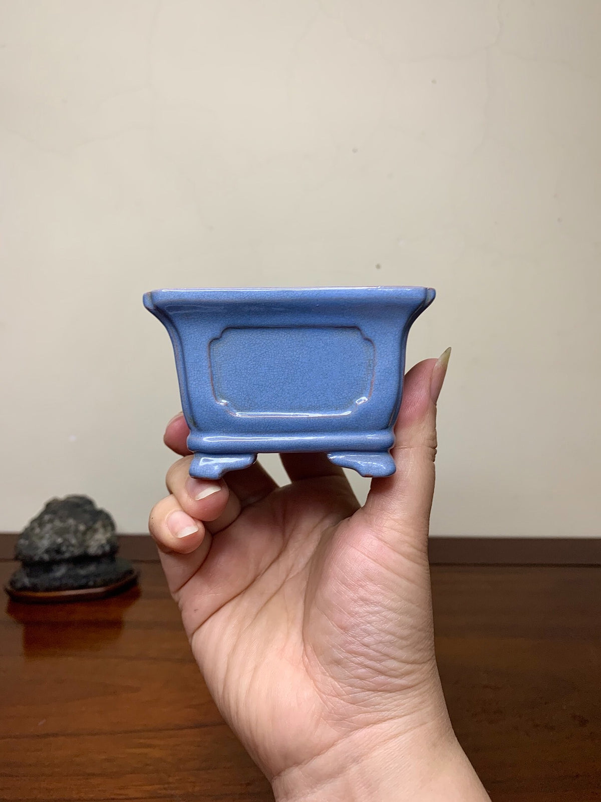 A hand holds a small, matte blue rectangular pot with a recessed center panel and four feet.