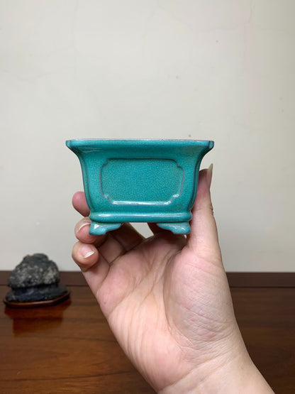 A hand holds a small, turquoise glazed rectangular pot with a textured surface and recessed panel.