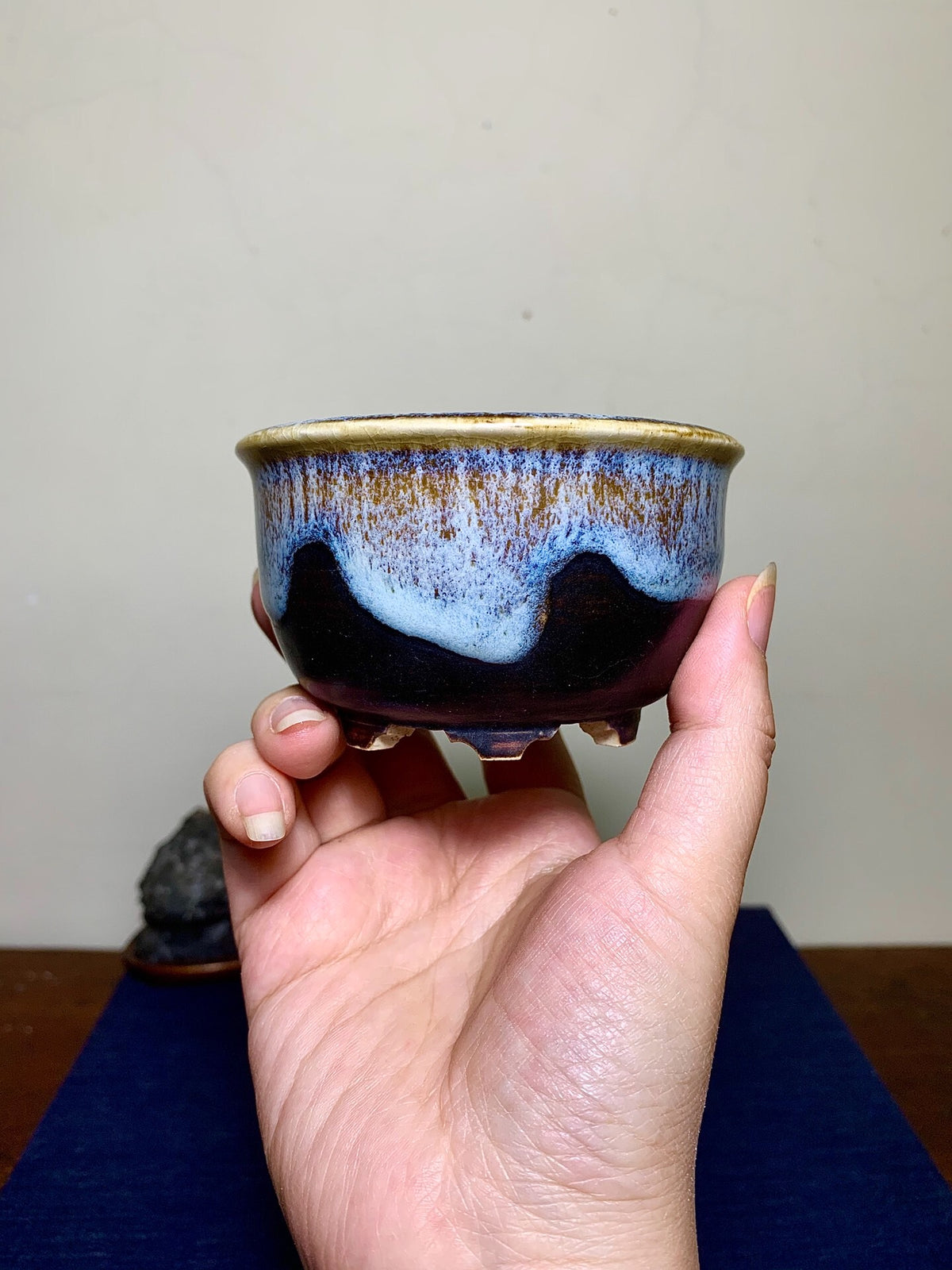 A hand holds a small, glazed ceramic pot with a dark base, blue and gold glaze, and three feet.