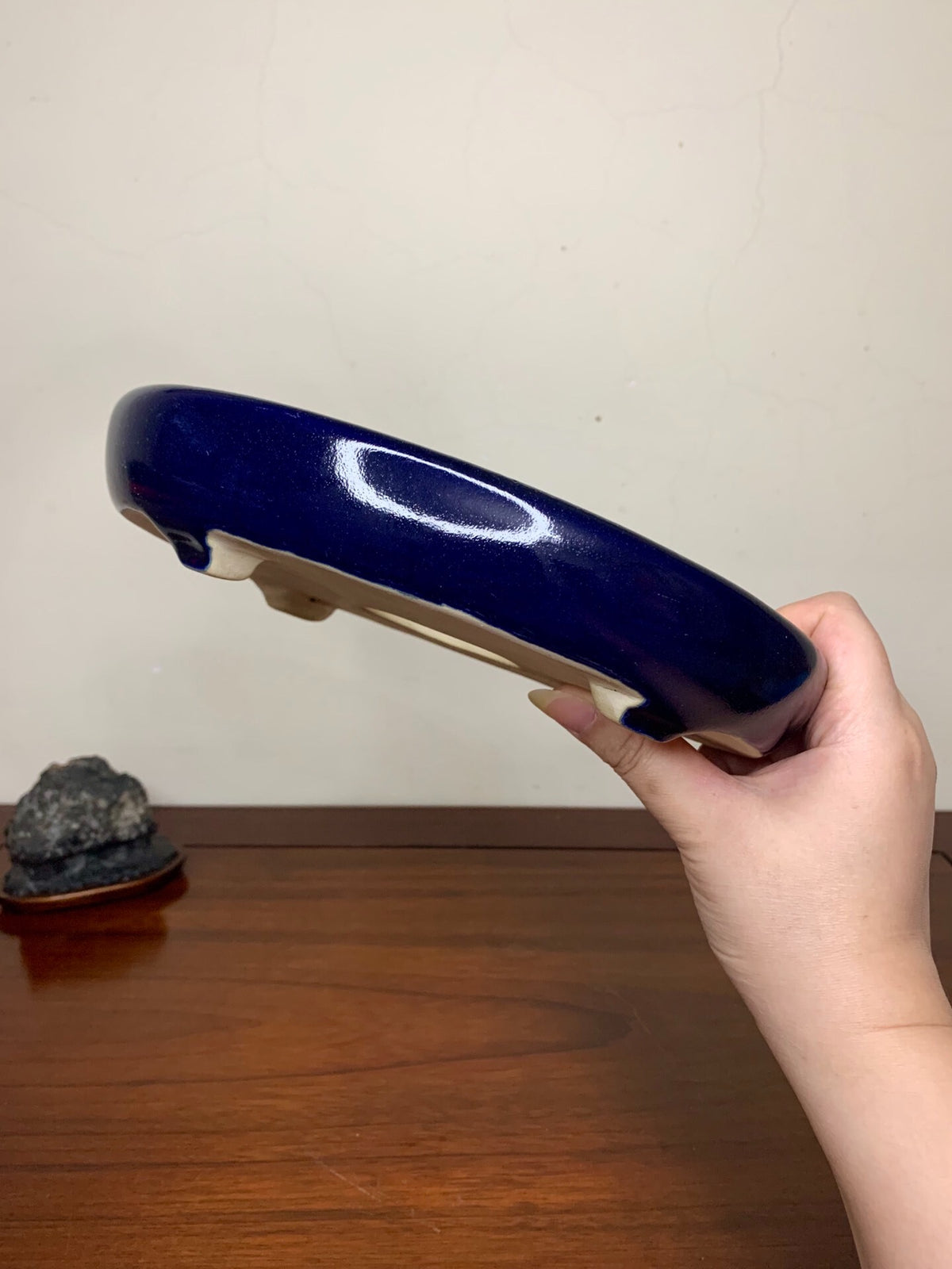 A hand holds a glossy, deep blue ceramic pot with a white oval mark on its curved rim.