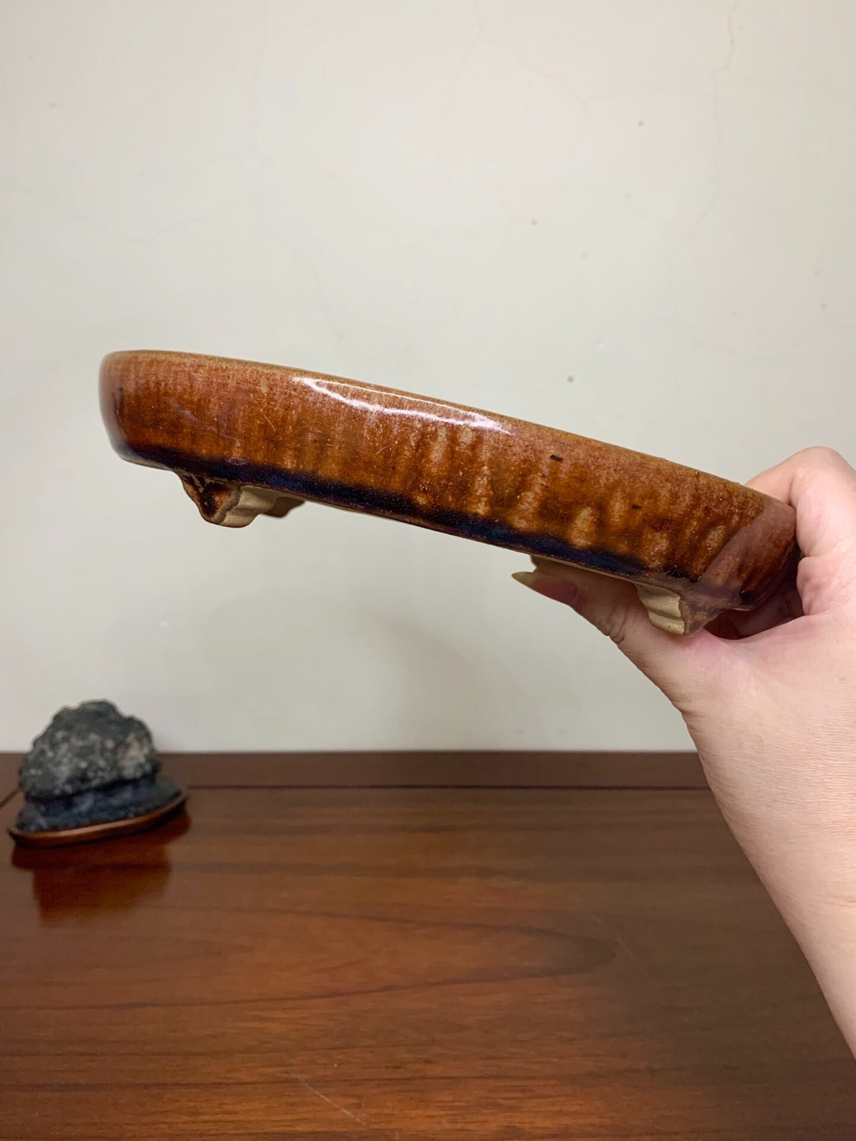 A hand holds a glossy, brown-glazed ceramic pot with a wide, shallow rim and two small feet.