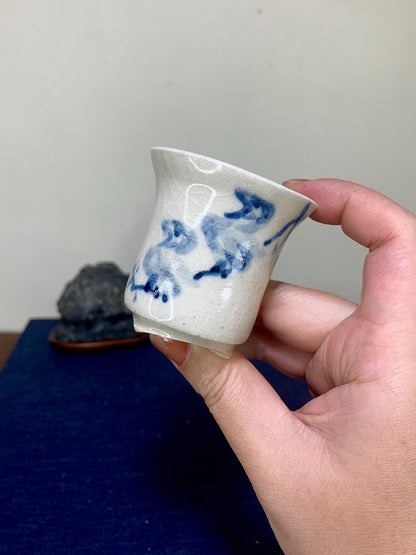 A hand holds a small, off-white ceramic cup with blue brushstroke patterns and a glossy finish.