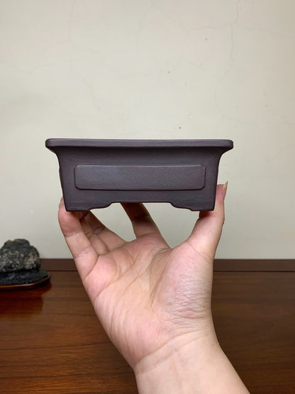 A hand holds a small, dark brown, rectangular unglazed ceramic pot with a recessed panel.