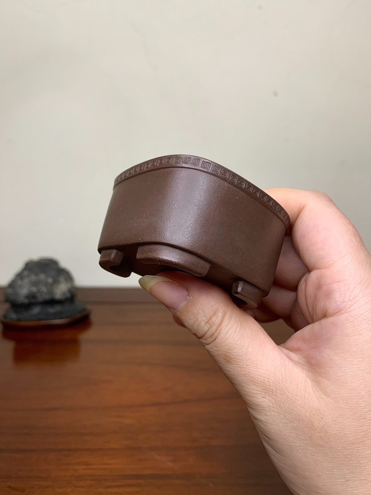 A hand holds a small, brown, matte-finished ceramic pot with a decorative rim and three feet.