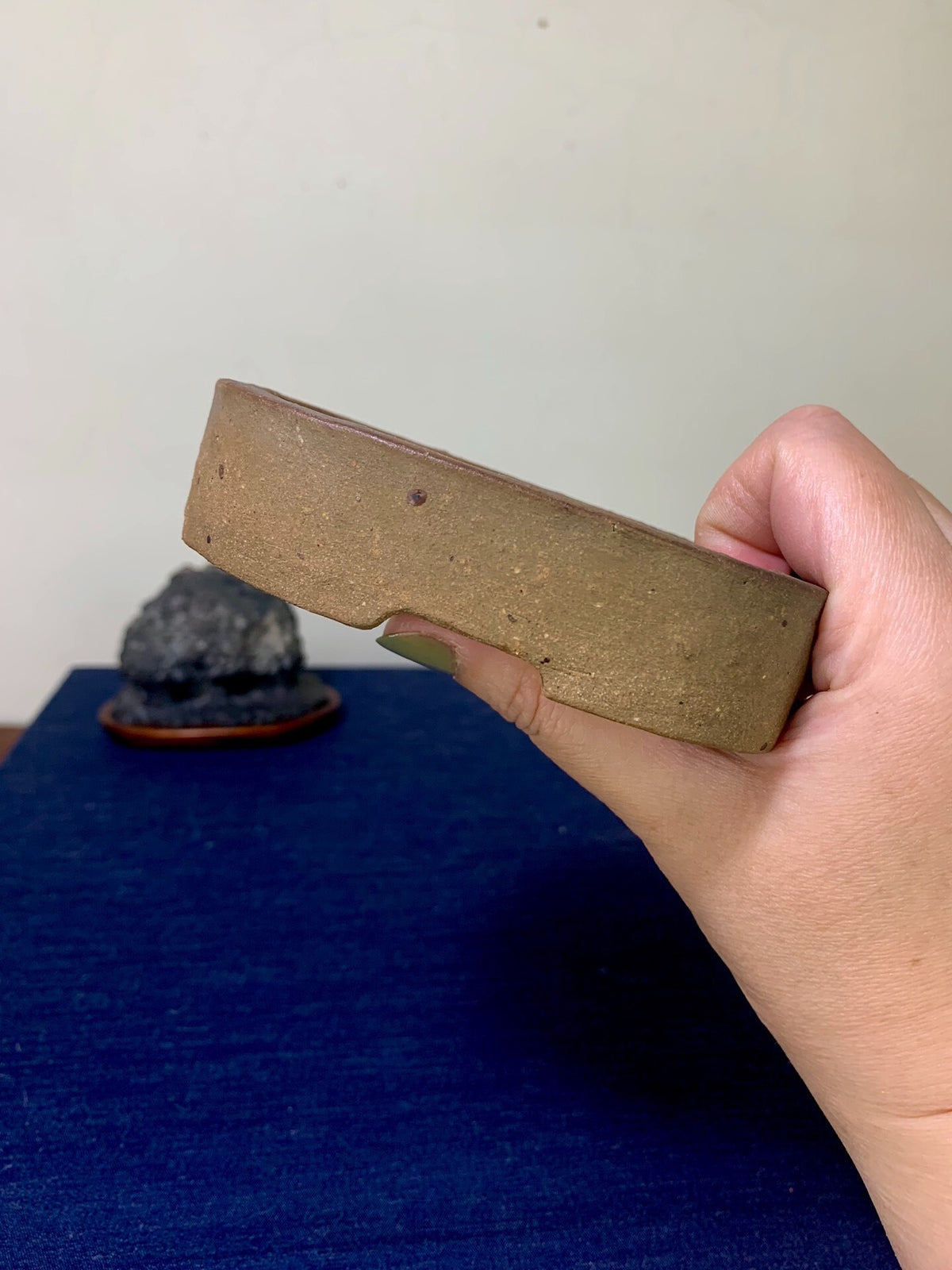 A hand holds a small, unglazed, brownish-grey rectangular pot with a rough, speckled surface.