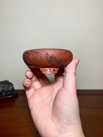 A hand holds a small, reddish-brown ceramic pot with carved mountain and tree scenes.
