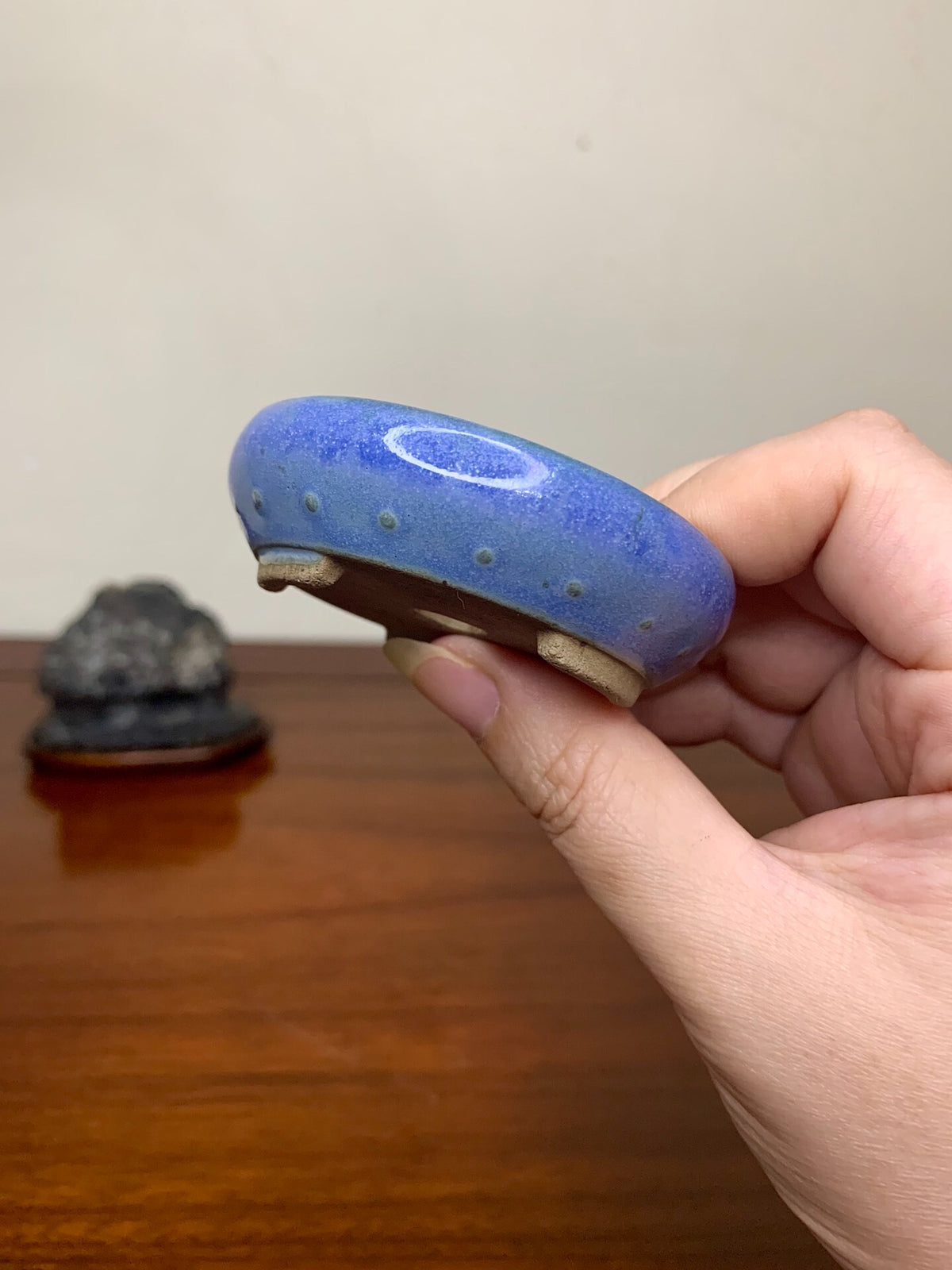 A hand holds a small, blue glazed pot with a white oval mark and raised dots.