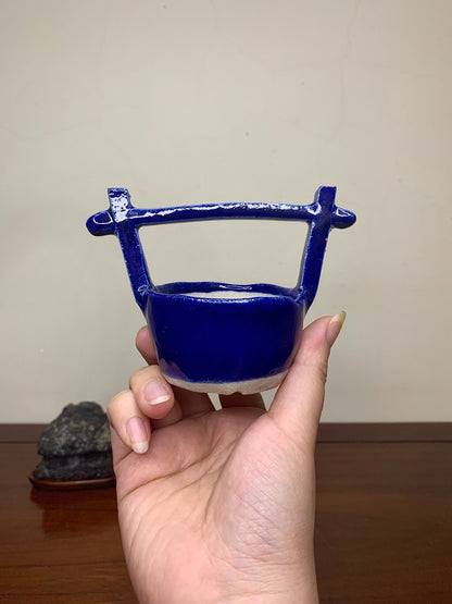 A hand holds a small, glossy cobalt blue ceramic pot with a handle and white base.