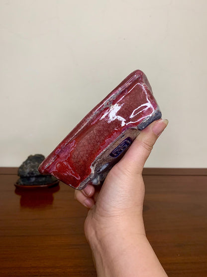 A hand holds a glossy, deep red glazed rectangular pot with a blue mark on its base.