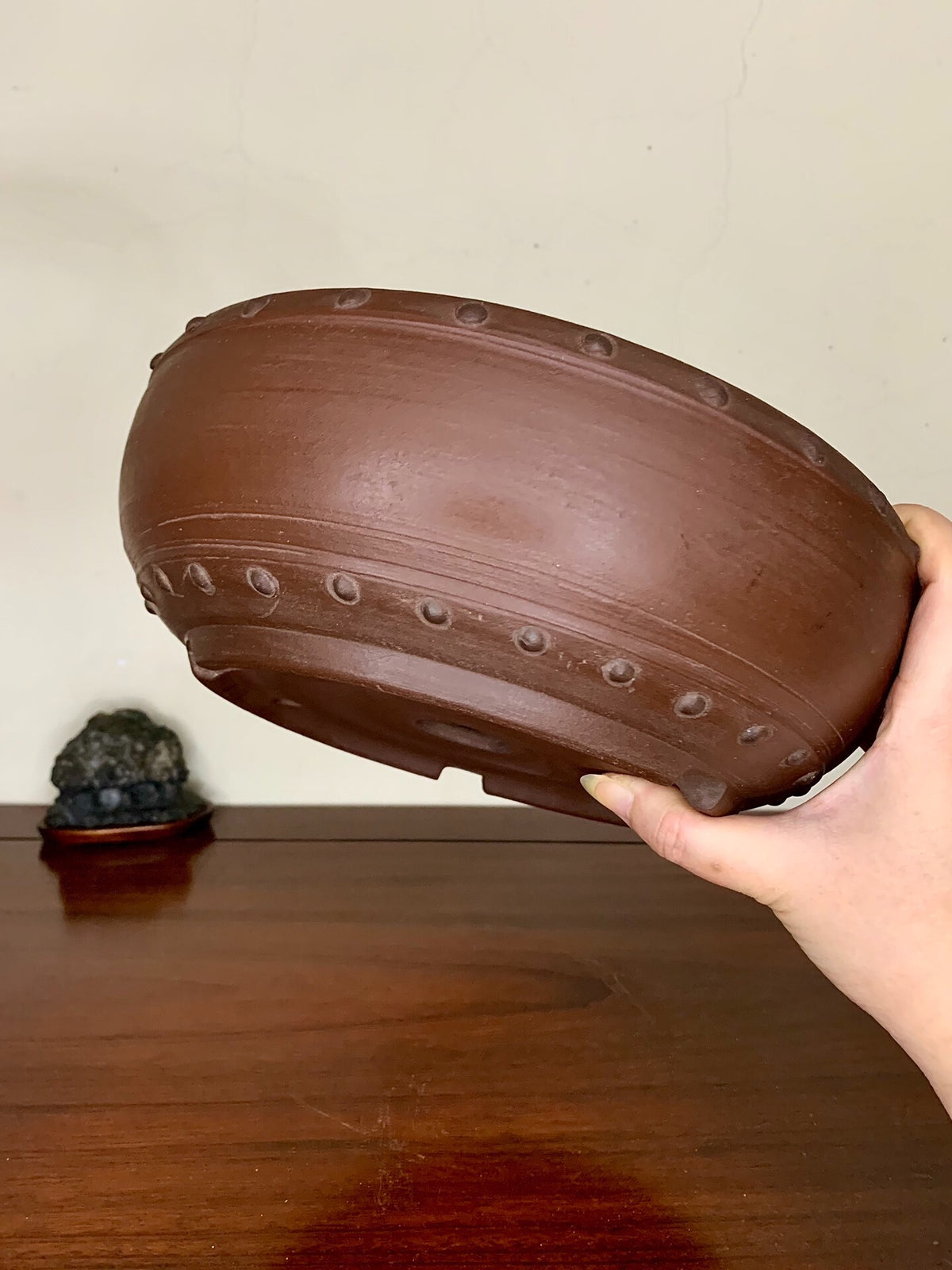 A hand holds a brown, unglazed ceramic pot with raised circular ridges around its rim.